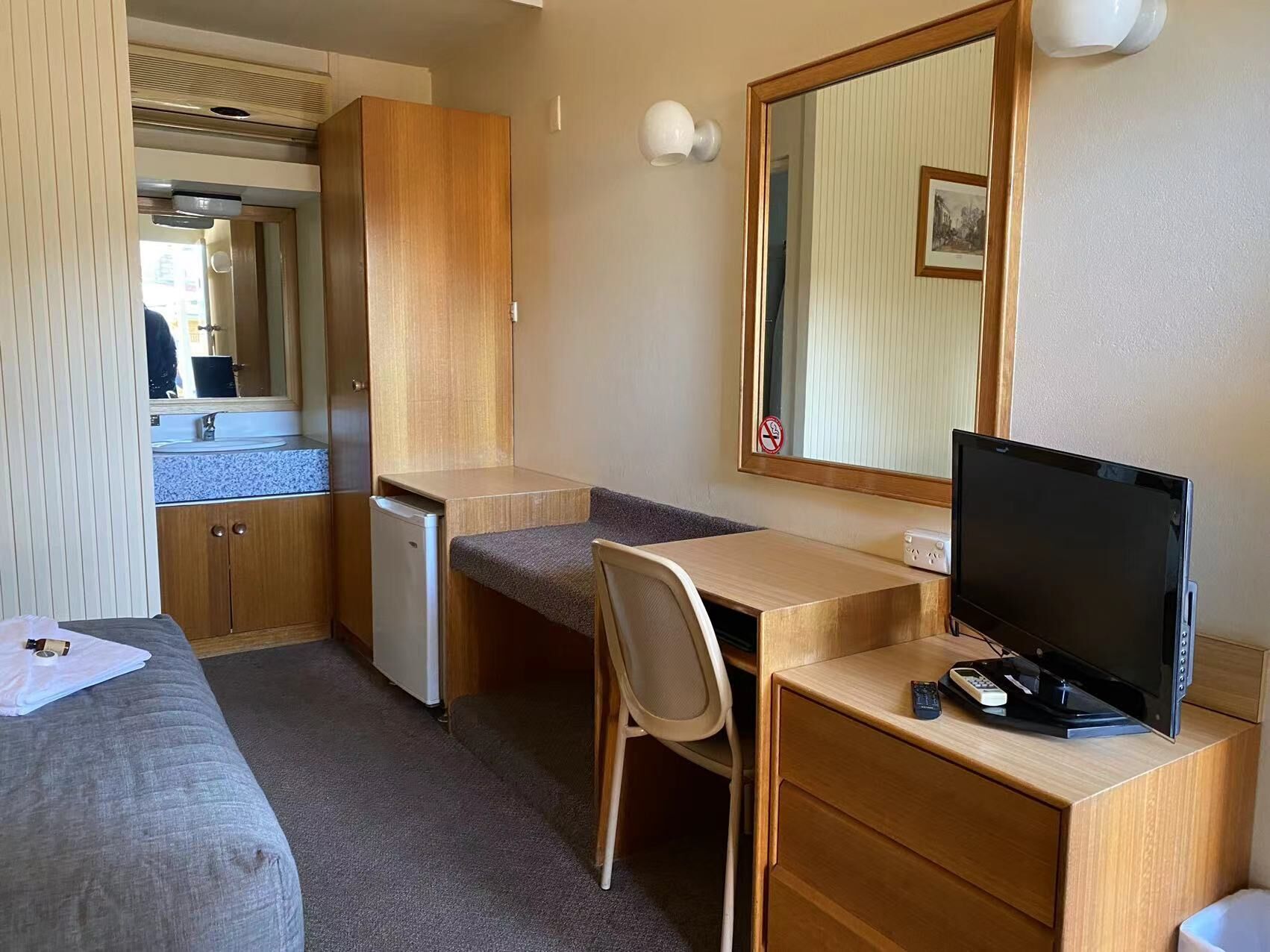 A compact hotel room with a kitchenette, a single bed, a desk with a chair, a television, and a large mirror.