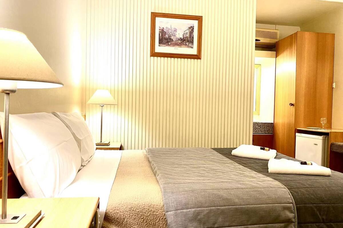 Ein Hotelzimmer mit Bett, Nachttisch, Lampe, einem gerahmten Bild an der Wand und einem Kleiderschrank.