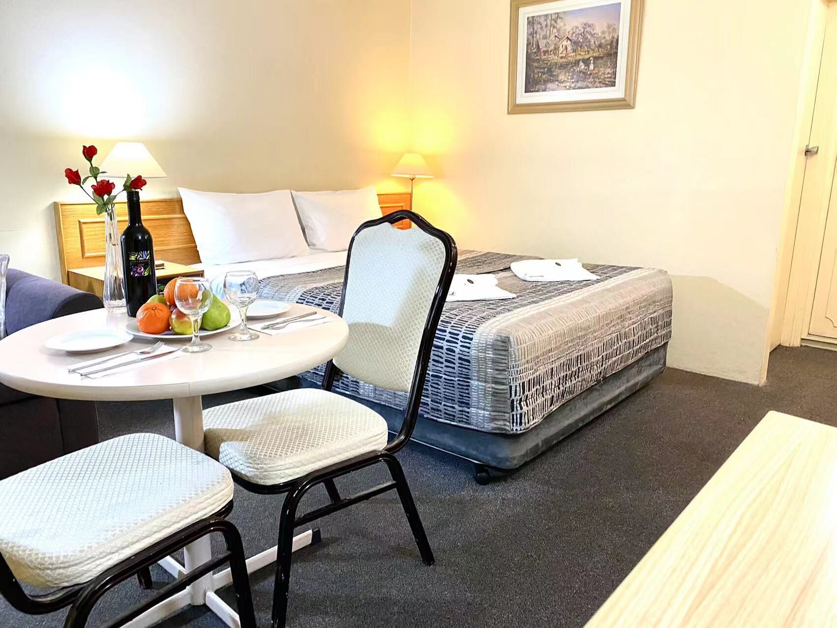 A small, cozy hotel room with a single bed, a dining table with chairs, a lamp, and a framed picture on the wall.