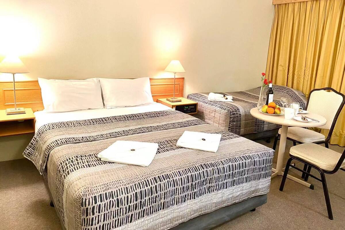 Ein Hotelzimmer mit einem großen Bett, zwei Nachttischen inklusive Lampen, einem kleinen runden Tisch mit Stuhl und einem Fenster mit Vorhängen.