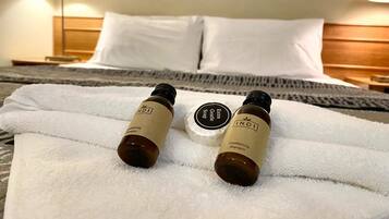 Une chambre d’hôtel avec un lit bien fait, une serviette blanche et deux flacons de shampoing et d’après-shampoing de la marque INDI posés sur le lit.