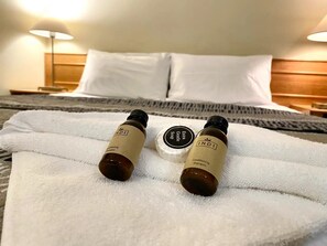 A hotel room with a neatly made bed, a white towel, and two bottles of INDI shampoo and conditioner on the bed.