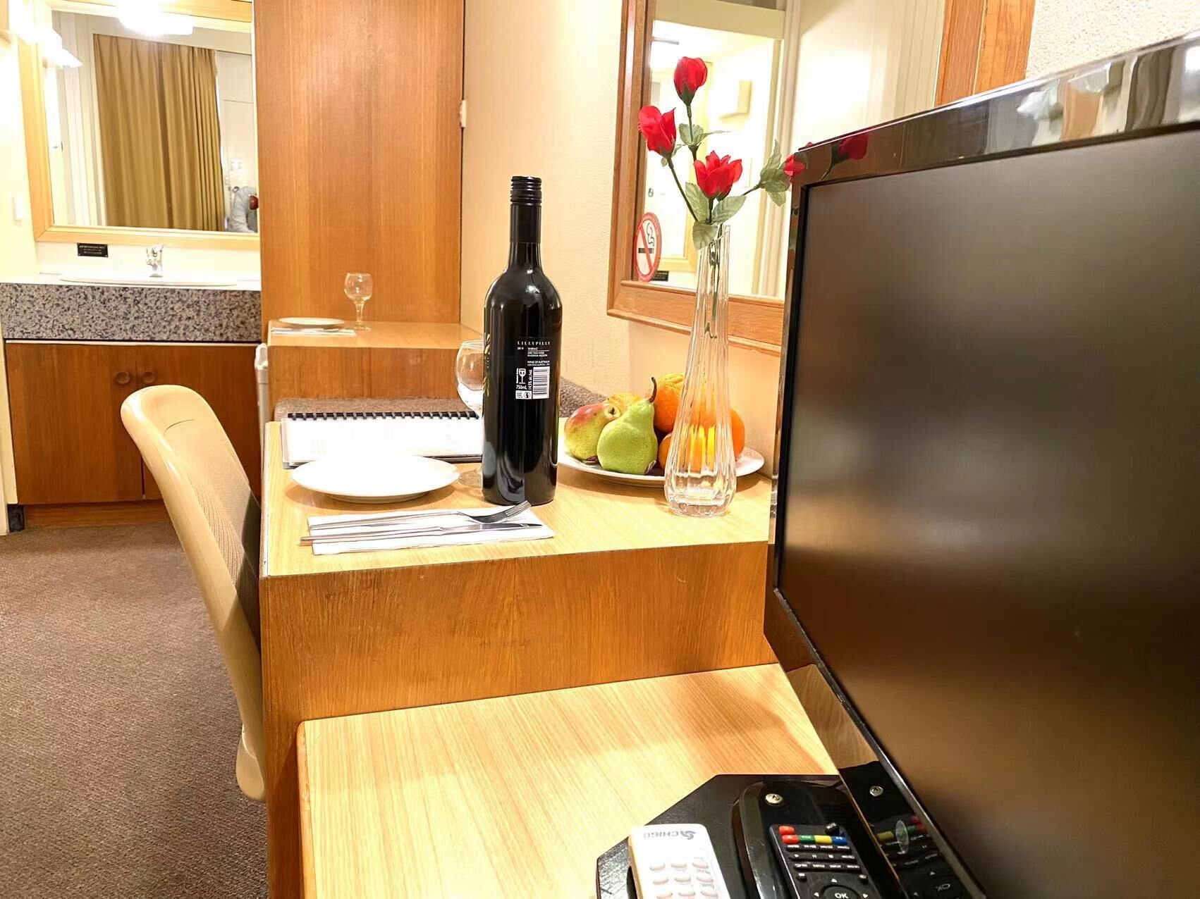 Habitación de hotel con un escritorio de madera, una laptop, una botella de vino, un florero con flores y una bandeja de fruta.