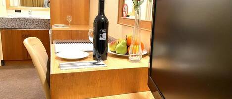 Habitación de hotel con un escritorio de madera, una laptop, una botella de vino, un florero con flores y una bandeja de fruta.