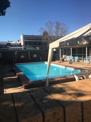 A swimming pool with a covered patio area, a fenced yard, and a building in the background.