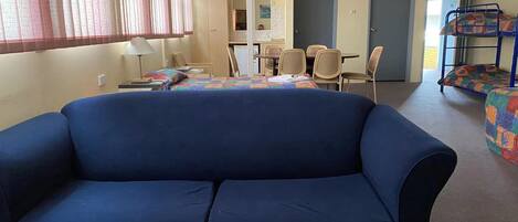 A room with a blue sofa, a dining table with chairs, a bunk bed, and a clock on the wall.
