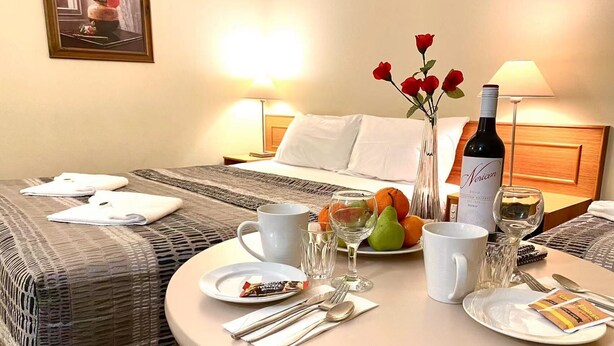 Une chambre d’hôtel avec un lit parfaitement fait, une table ronde dressée pour le dîner, avec des assiettes, des verres, des couverts et une bouteille de vin.