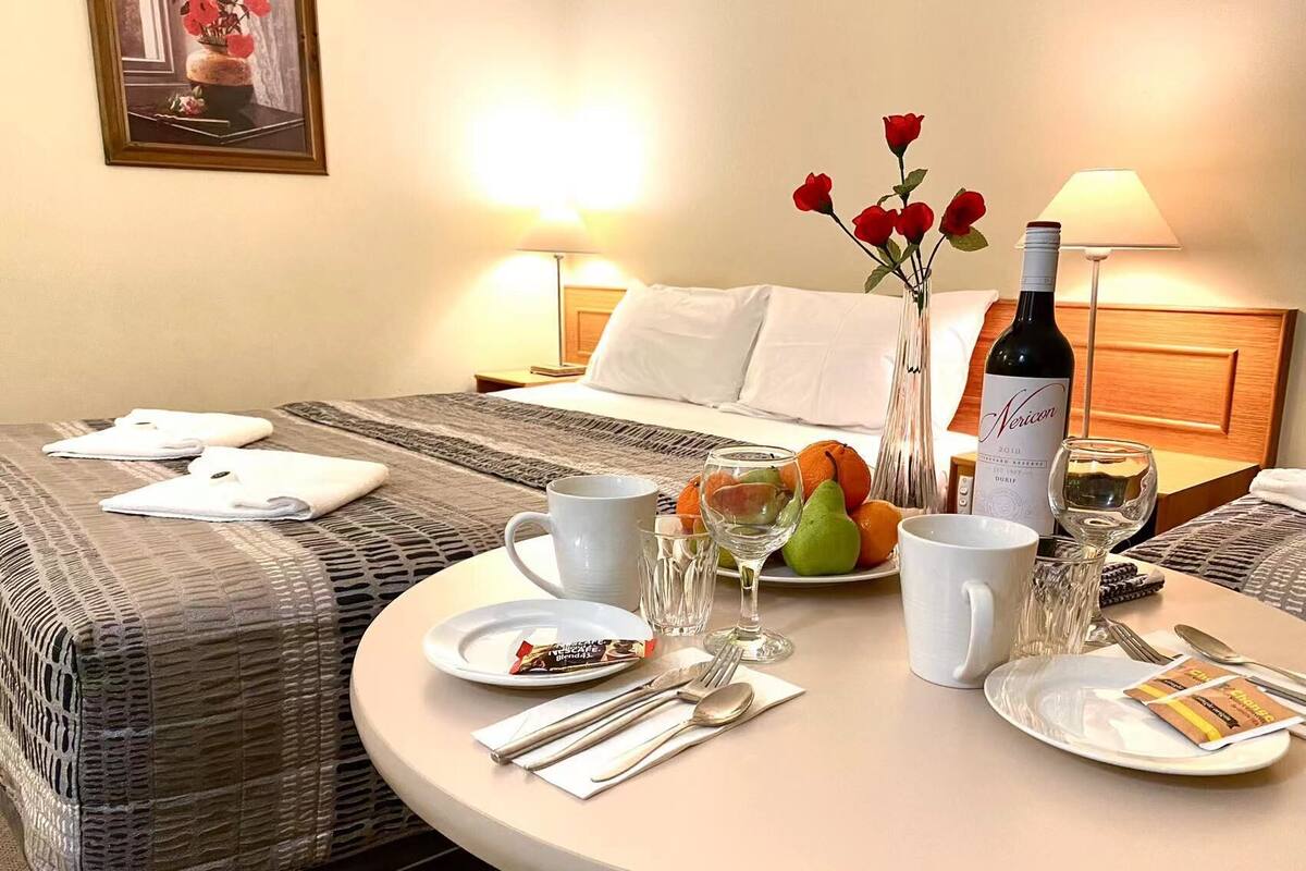 Ein Hotelzimmer mit einem ordentlich bezogenen Bett, einem runden Esstisch gedeckt mit Tellern, Gläsern, Besteck und einer Weinflasche.