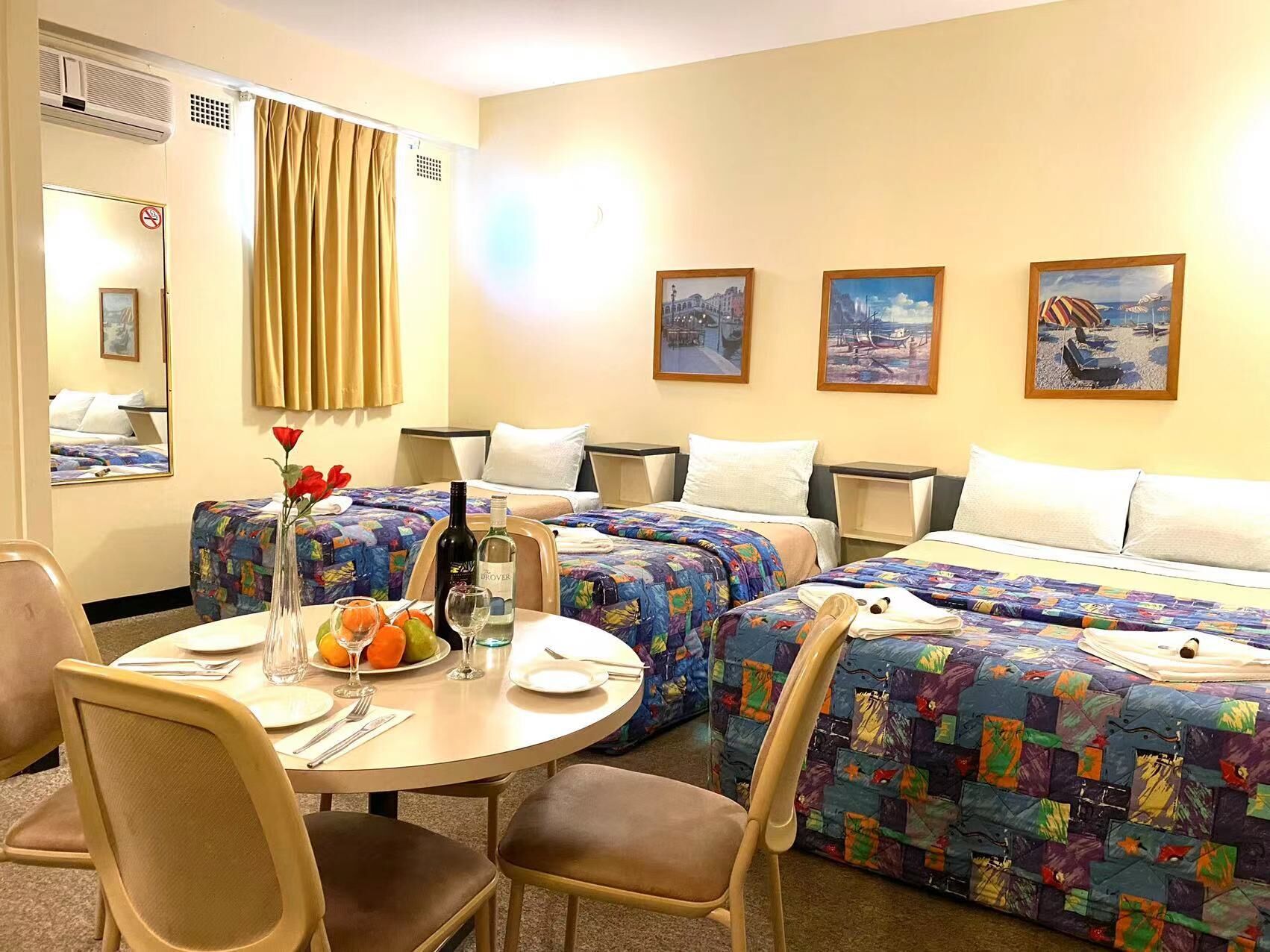 A hotel room with two beds, a dining table with chairs, and a small table with a bottle and glasses.