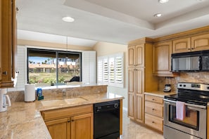 A kitchen with wooden cabinets, a dishwasher, an oven, and a window with blinds. - Refreshed Waterfront Desert Escape end unit on Golf Course facing West (Palm Desert)