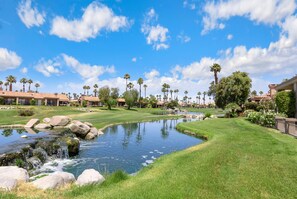 A golf course with a pond, palm trees, and buildings in the background. - Waterfront Desert Escape, Waterfalls, Membership Access (Palm Desert)