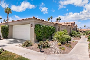 A residential house with a garage, a well-maintained garden with various plants and flowers, and a clear blue sky. - Refreshed Waterfront Desert Escape end unit on Golf Course facing West (Palm Desert)