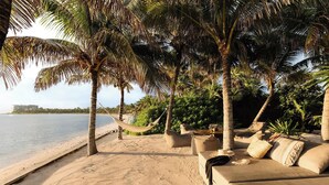 Um espaço de descanso à beira-mar com uma rede, palmeiras e uma praia arenosa.