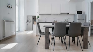 A modern kitchen with white cabinetry, a dining area with a table and chairs, and a visible refrigerator. - Nilo – Historic Apartment with Patio by Casa N (Napoli)