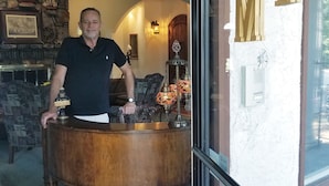 A man standing behind a wooden bar counter with a stone fireplace in the background. - Tuscan Manor (Eureka Springs)