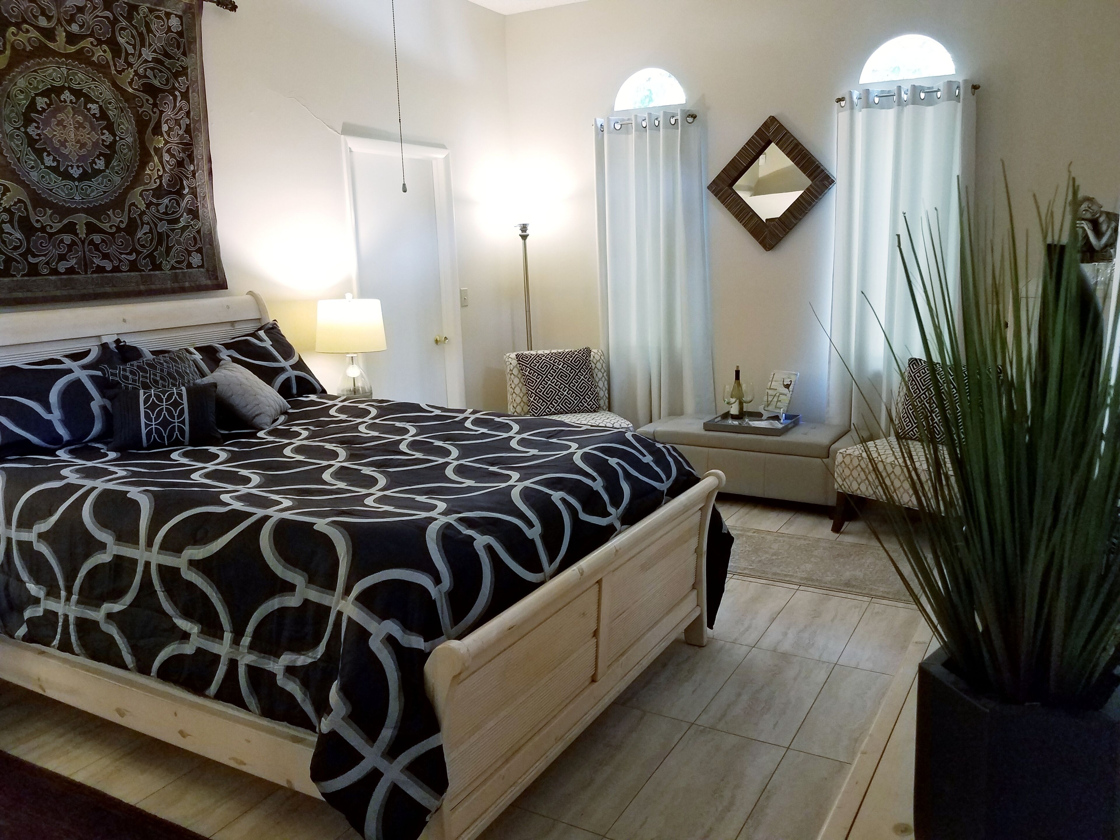 A bedroom with a large bed, a bedside table with a lamp, a chair, a mirror, and a potted plant.