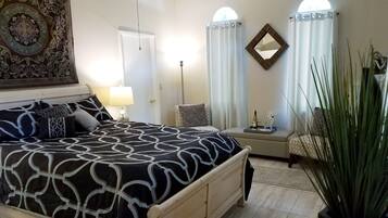 A bedroom with a large bed, a bedside table with a lamp, a chair, a mirror, and a potted plant.
