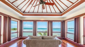 Une pièce dotée d’un ventilateur de plafond, de grandes fenêtres donnant sur l’océan, d’un plancher en bois et d’un canapé beige.