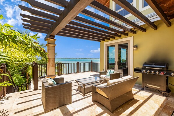 A covered patio with wicker furniture, a grill, and a view of the sea.