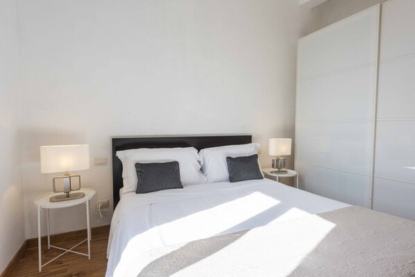 A modern hotel room with a bed, two bedside tables with lamps, a large wardrobe, and a wooden floor.