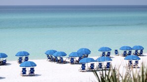 Een strand met rijen blauwe strandstoelen en parasols, een heldere blauwe zee en een zandstrand.