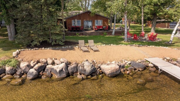 A cabin with a deck, surrounded by a sandy area and a small pond. - Premier Location on Crosslake - Hot Tub - Pontoon Rental - Waterslide - Games  (Crosslake)