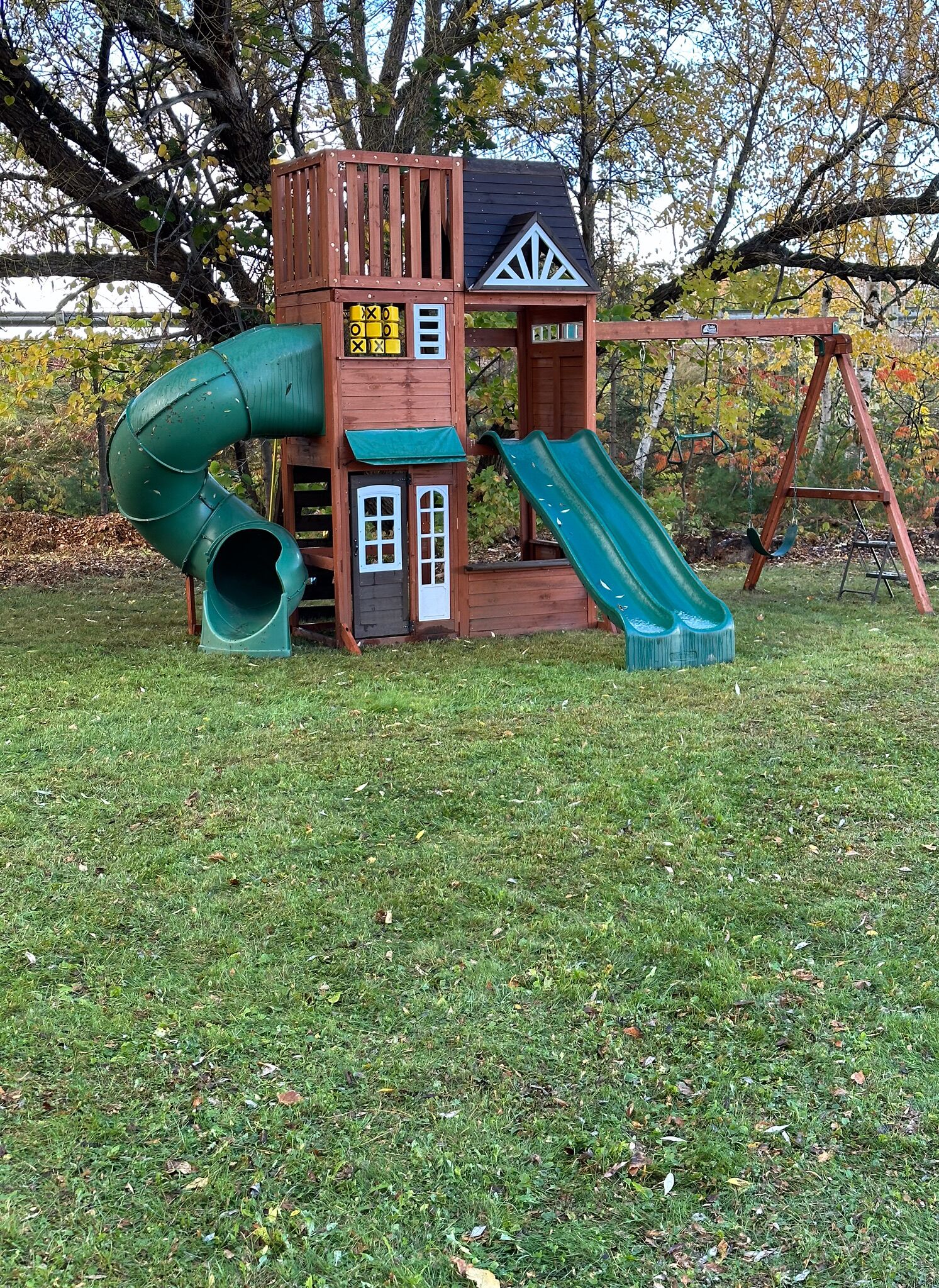 A wooden playhouse with a slide and a swing set in a grassy yard.