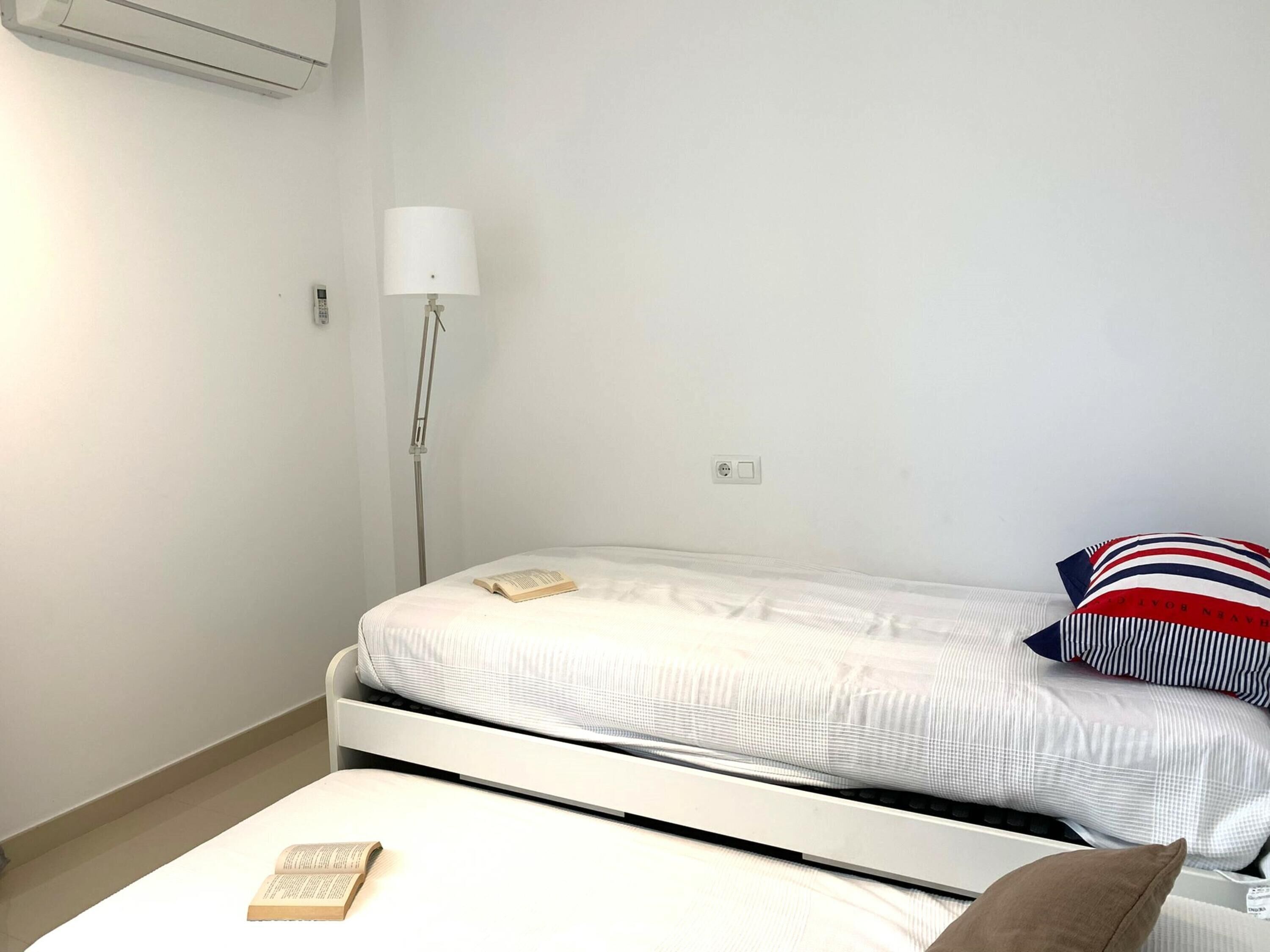 Een kleine, schone kamer met een enkel bed, nachtkastje, lamp en een wandairconditioner.