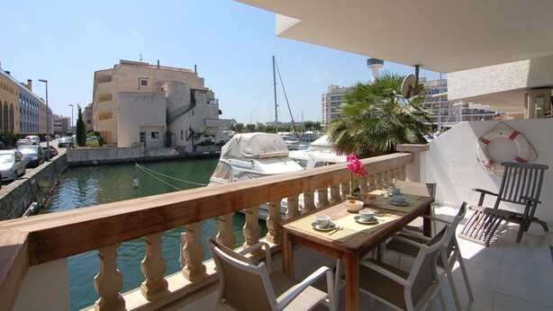 A balcony with a wooden railing overlooking a marina with boats, a palm tree, and buildings in the background. - Charming apartment for rent in Empuriabrava, Costa Brava. 1100m from the beach, capacity for 4 people. (Empuriabrava)