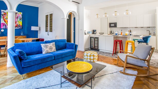 A modern living room with a blue sofa, a glass coffee table, and a kitchen area with white cabinets and a red stool. - Appartement Armény - Welkeys (Marseille)