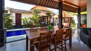 A covered outdoor dining area with a pool, wooden furniture, and tropical plants.