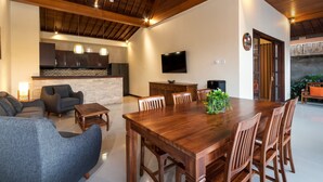 A spacious living area with a wooden dining table, comfortable seating, a flat-screen TV, and a kitchen in the background.