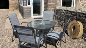 Un patio al aire libre con una mesa de vidrio redonda y cuatro sillas, un muro de piedra y una puerta de madera.