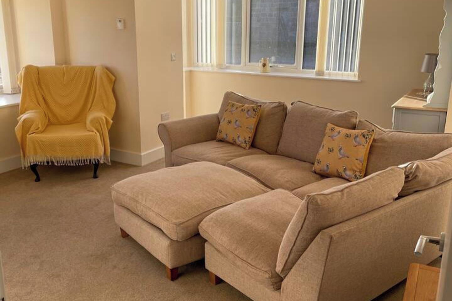 A living room with a beige sectional sofa, a yellow armchair, and a window with blinds.