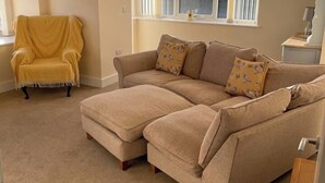 Una sala de estar con un sofá seccional color beige, un sillón amarillo y una ventana con persianas.