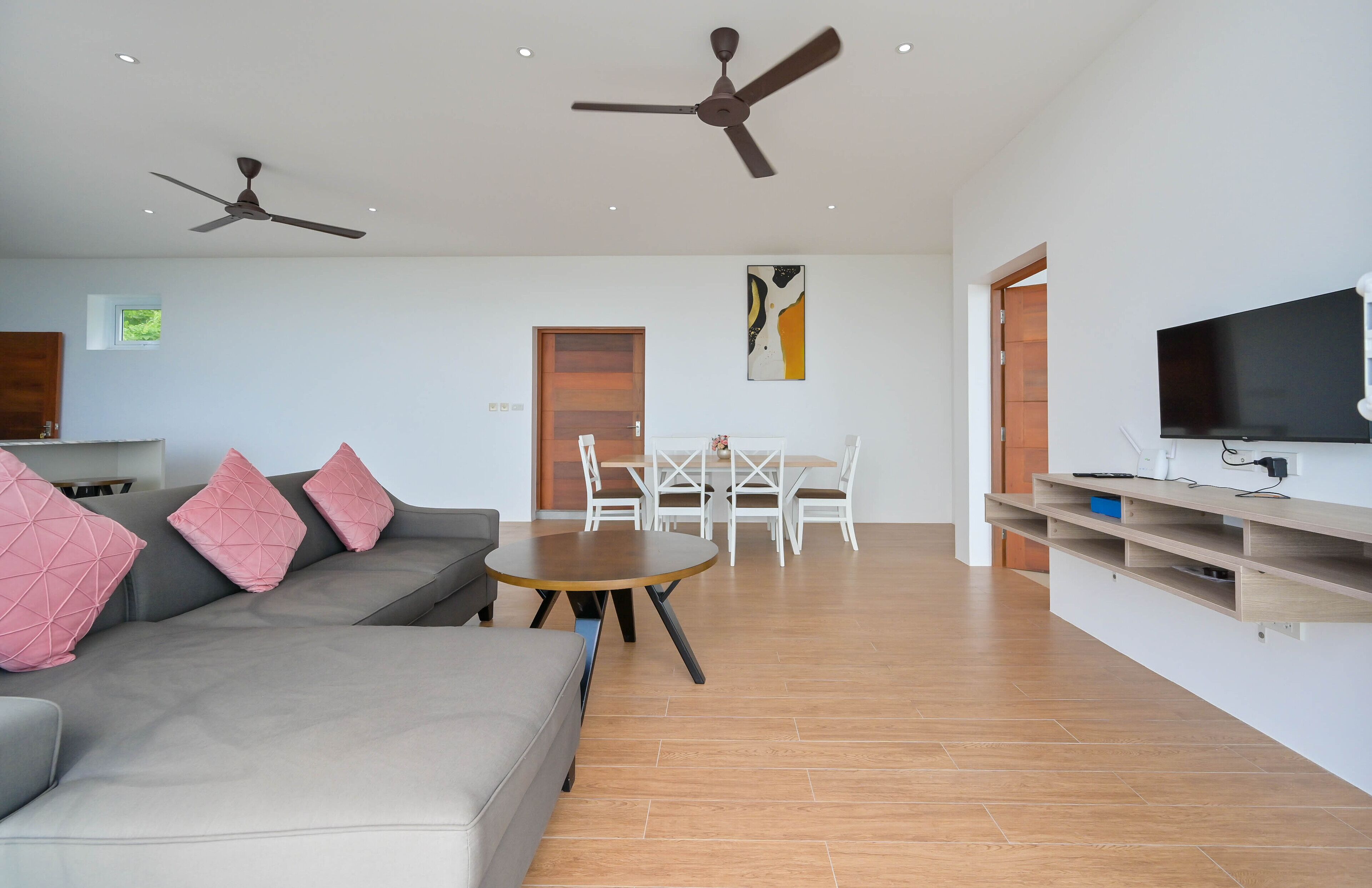 A modern living room with a grey sofa, pink cushions, a round wooden coffee table, a dining table with white chairs, ceiling fans, a flat-screen TV, and wooden flooring.