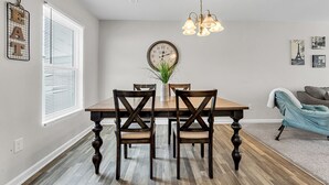 A dining area with a wooden table, four chairs, a hanging light fixture, a wall clock, and a window with blinds. - Sleeps 6 - 3 Queen Beds - 2 Mi Wake Forest + Hospital (Winston-Salem)