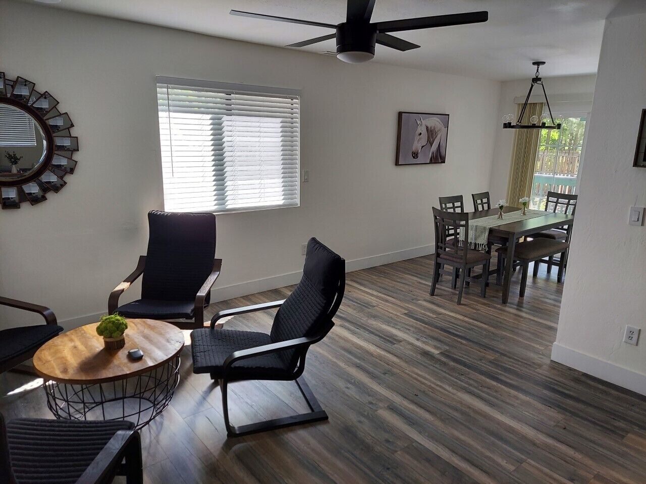 A spacious living room with a dining area, wooden flooring, a ceiling fan, and a large mirror on the wall.