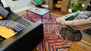 A living room with a black leather sofa, a colorful rug, a fireplace, and a television mounted above it.