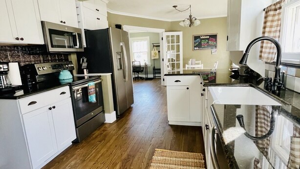 A kitchen with white cabinets, a black refrigerator, a microwave, and a stove. There is a woven mat on the floor and a hanging light fixture above the sink.