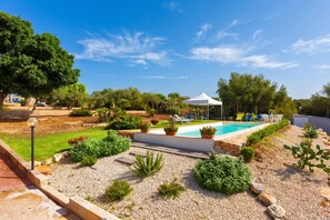 A pool area with a white tent, surrounded by gravel and plants, with trees in the background.