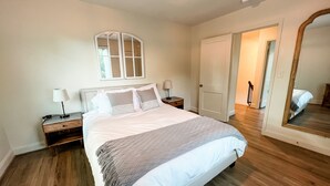 Un dormitorio con una cama grande, una mesita de noche, una lámpara, un espejo y una puerta que da a otra habitación.