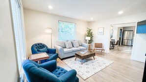 Una sala de estar con un sillón azul, un sofá gris y una mesa de centro de madera. Se ve una ventana con persianas, una lámpara y una planta.