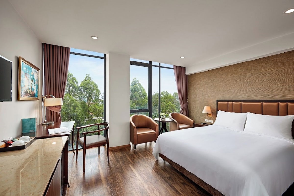 Habitación de hotel con cama, escritorio, dos sillas, mesa y ventanas amplias que dan a vistas de árboles.