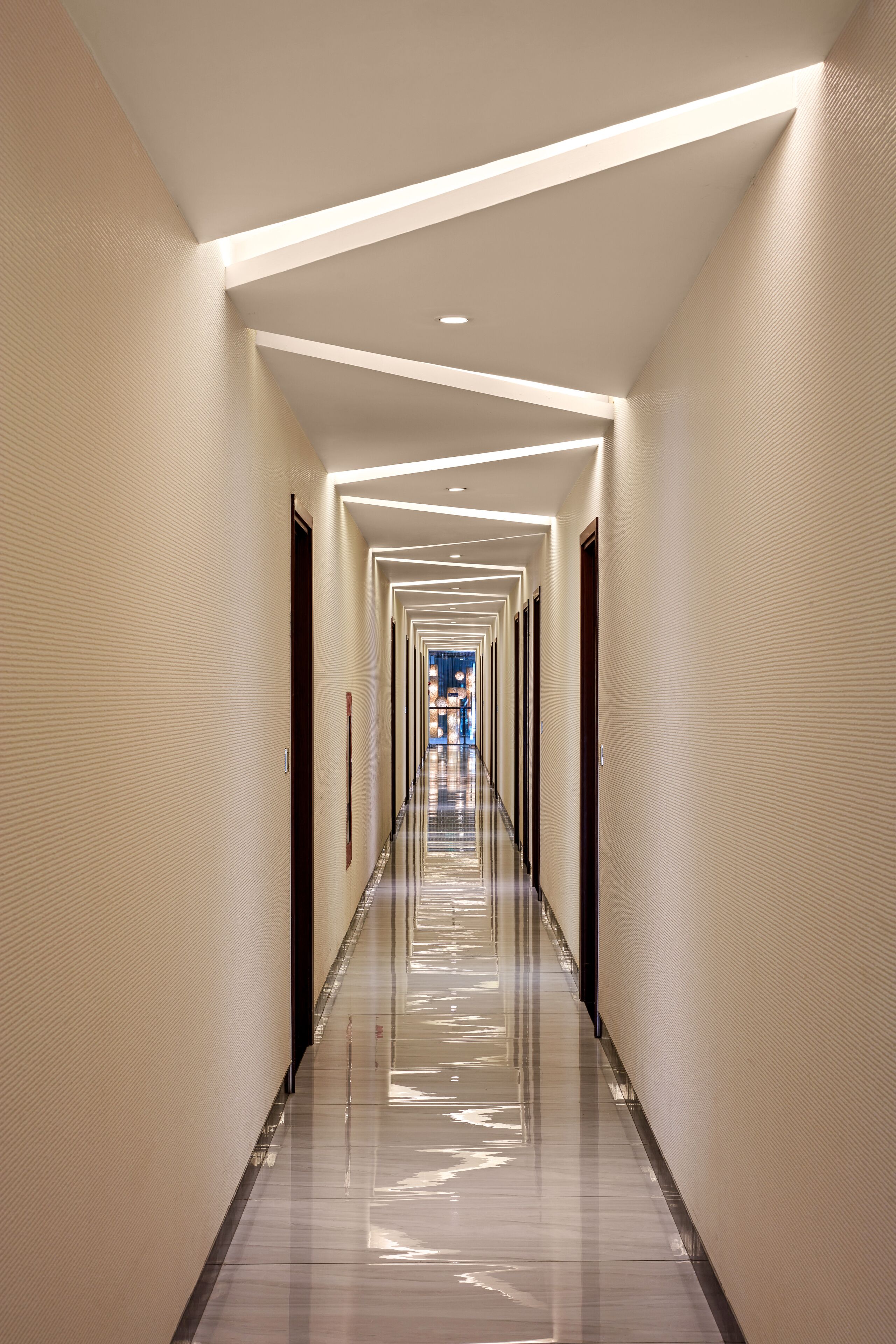 Un largo y estrecho pasillo de hotel con suelo reflectante y puertas distribuidas uniformemente a ambos lados.
