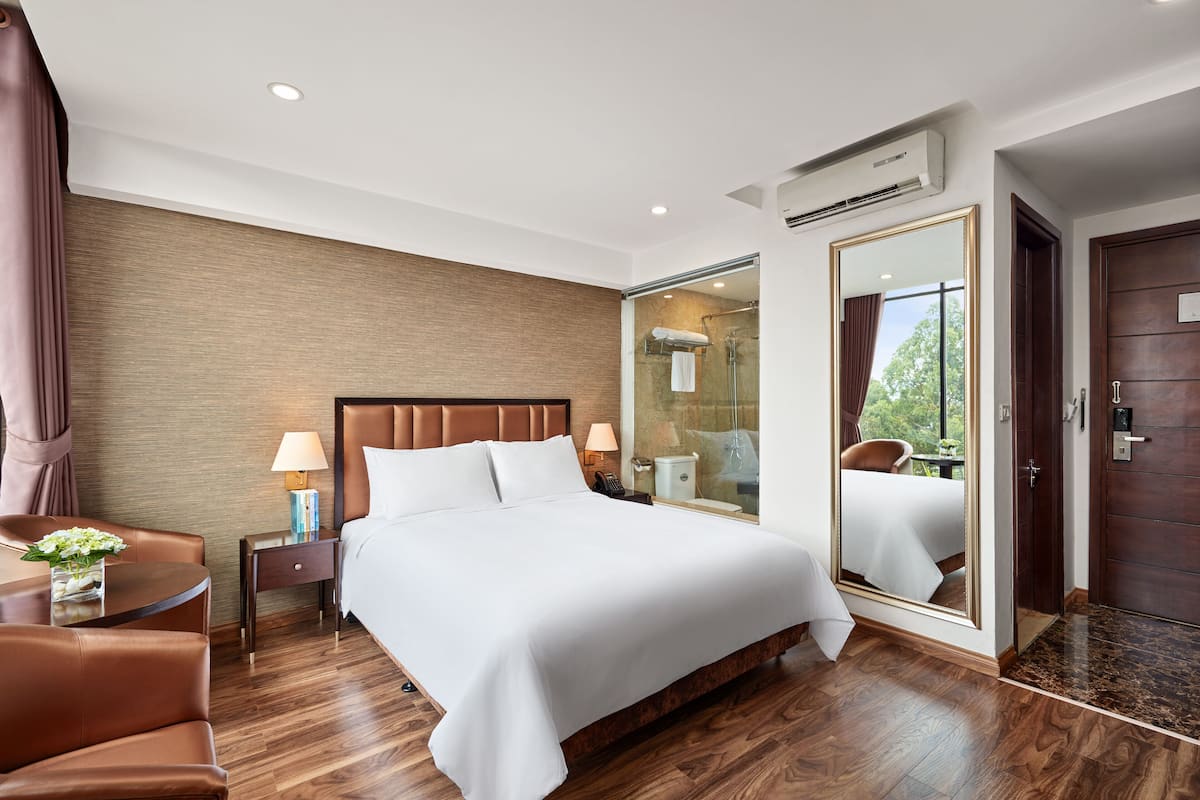 A hotel room with a large bed, a sitting area with a chair, a small table with a vase, a nightstand, a mirror, and a door leading to a bathroom.