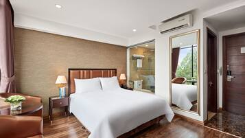 Habitación de hotel con una cama grande, un espacio de descanso con una silla, una mesita con un florero, una mesita de noche, un espejo y una puerta que da a un baño.