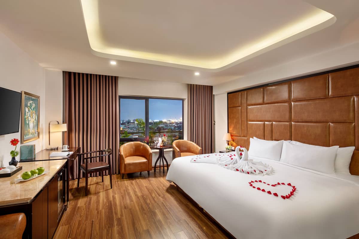 A hotel room with a large bed, a desk with a chair, a small table, and a view of the city.