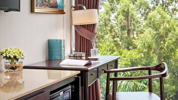 A room with a desk, chair, TV, painting, curtains, and a view of trees.