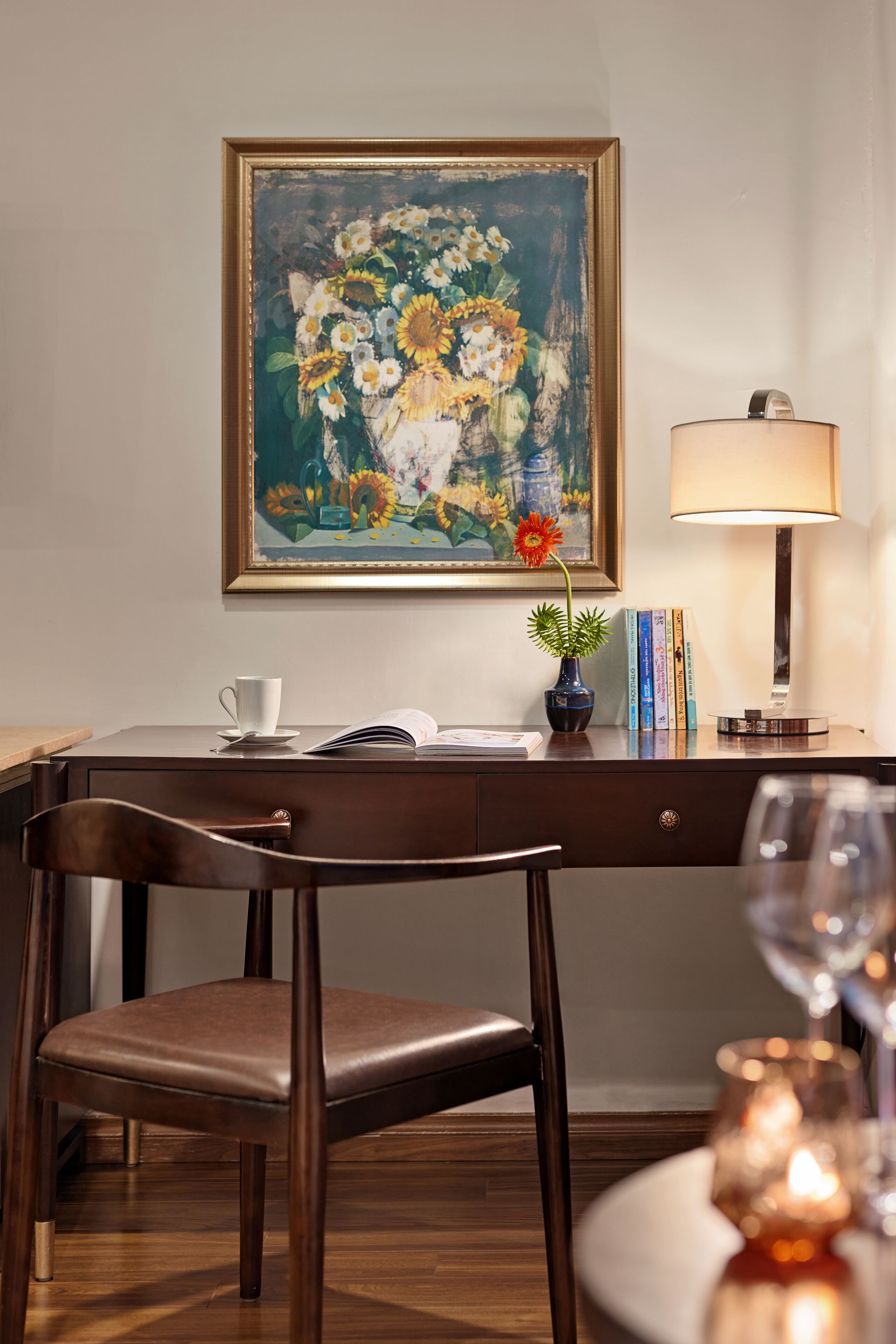 Une pièce comprenant un bureau, une chaise et une table avec un verre à vin. Il y a un tableau au mur et une lampe sur le bureau.